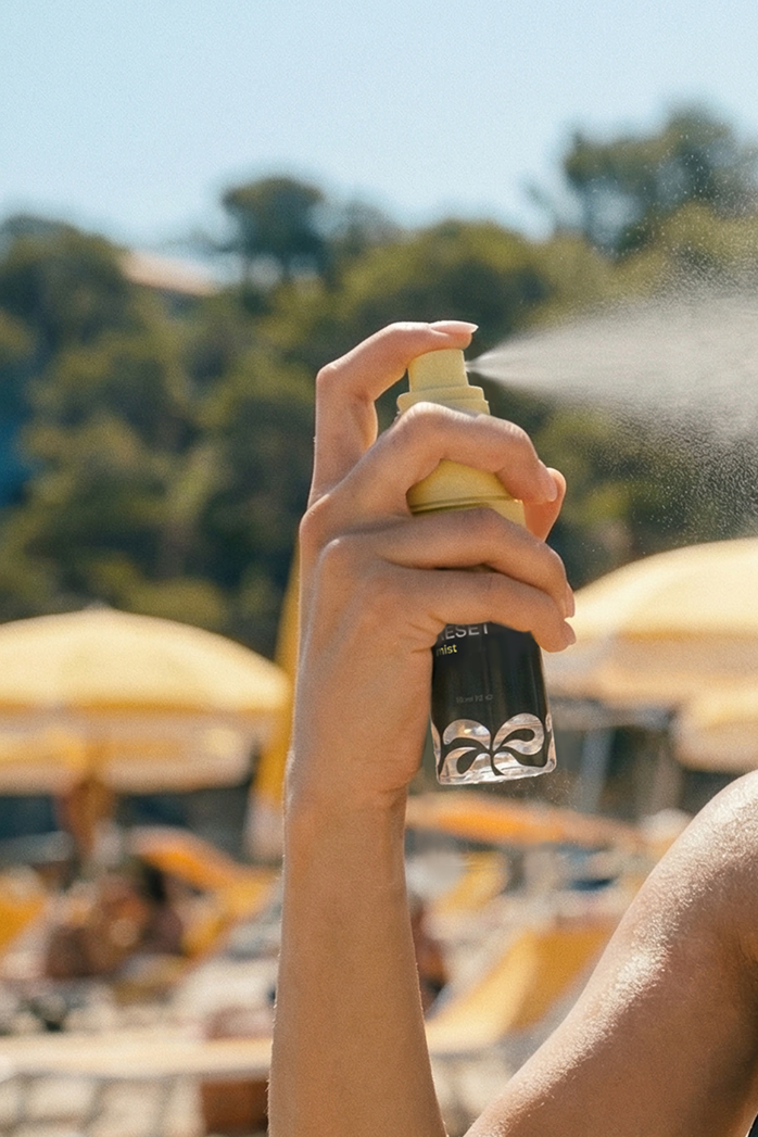 Hand holding a mist spray bottle outdoors, 
fine mist dispersing in the air against 
a bright sky with yellow beach umbrellas below.