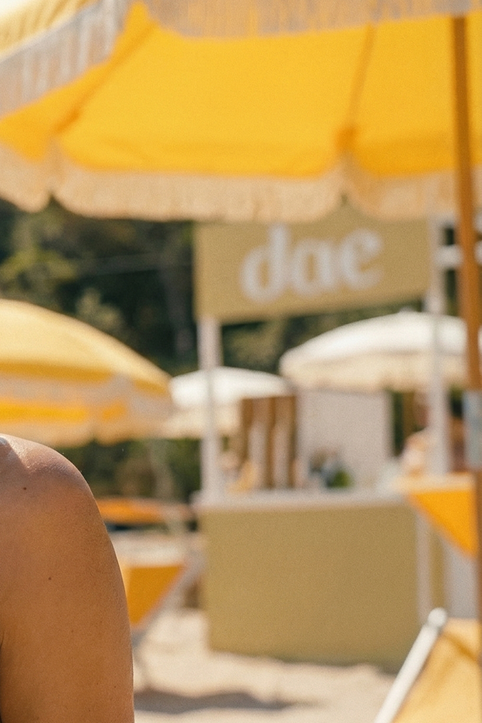 Sun-drenched beach scene with yellow 
striped umbrellas and a bare shoulder 
in the foreground, daezls signage visible 
in the background.